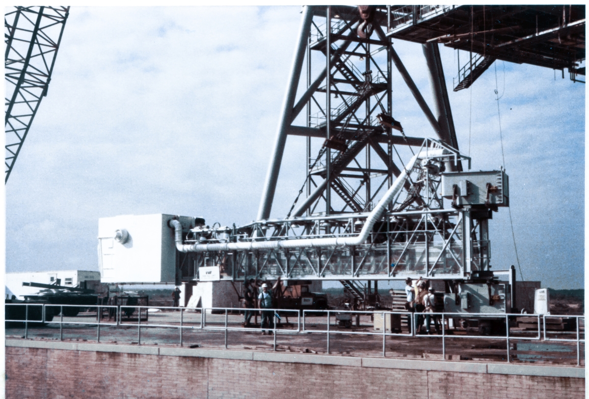 Image 132. OAA Lift 4 of 9. This is the last image taken of the OAA before it was lifted up to its final location and bolted to its strongback on the side of the FSS at Space Shuttle Launch Complex 39-B, Kennedy Space Center, by Union Ironworkers working for Ivey Steel. It is still resting on its support stanchions (but you can also see that the lift rigging is near-fully tensioned, exerting significant upward force on the Arm, but not quite enough to cause it to break contact with the concrete it still rests upon), but Ivey's ironworkers are in the process of removing the bolts which attach the support stanchion to the Arm, closest to the Hinge Boxes. When it goes up from here, the stanchions will remain on the Pad Deck. Elsewhere, the opening in the Environmental Chamber which the Shuttle Crew passes through to access the hatch on the side of the Orbiter, has received its protective cover, and we'll get to see that in the next image in this series. We're very very close to lifting the Arm at this point. Photo by James MacLaren.